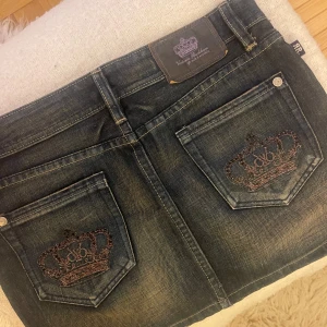 Victoria Beckham kjol - Snygg jeanskjol från Victoria Beckham i mörk tvätt med coola broderade kronor på bakfickorna. Kjolen har en klassisk femficksdesign och stängs med dragkedja och knapp.