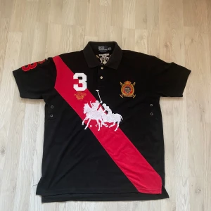 Svart pikétröja från Ralph Lauren - Säljer en snygg svart pikétröja från Ralph Lauren med en röd diagonal rand och broderade detaljer. Tröjan har korta ärmar och en klassisk passform. Perfekt för en stilren look! Passar som en M