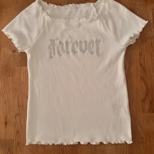 Vit ribbad topp från Lindex - Säljer en superfin vit ribbad topp från Lindex i storlek 146/152. Den har korta ärmar och texten 'Forever' i glittriga stenar på framsidan. Toppen är i ekologisk bomull och har en söt volangdetalj vid halsen och ärmsluten. Endast använd en gång 