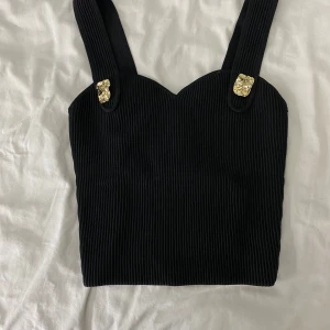 Svart ribbad topp med gulddetaljer - Snygg svart ribbad topp med breda axelband och guldfärgade knappliknande detaljer. Perfekt för en utekväll eller fest. Toppen har en tight passform och är i mycket bra skick.