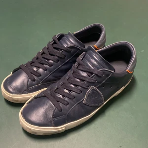 Philipe model - Säljer ett par mörkblå Philipe model PM/78EDT i skinn med snygga orange detaljer vid hälen. Skorna har en klassisk snörning och en vit sula som ger en cool kontrast. Perfekta för både vardag och fest! De är i bra skick med slitage som design på sulan. 