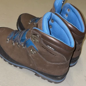 Bruna kängor från Everest - Säljer ett par robusta bruna kängor från Everest i mycket bra skick. De har blå detaljer och snörning, perfekt för höst- och vinteräventyr. Tillverkade i slitstarkt material med bekväm passform och bra grepp.