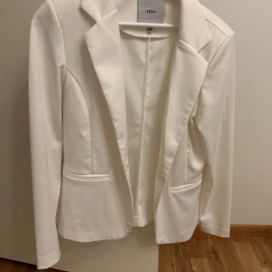 Vit kavaj - Snygg och stilren vit kavaj från ICHI i mycket bra skick. Perfekt för både vardag och fest. Den har en klassisk design med långa ärmar och två fickor framtill. Passar bra till både jeans och klänning.