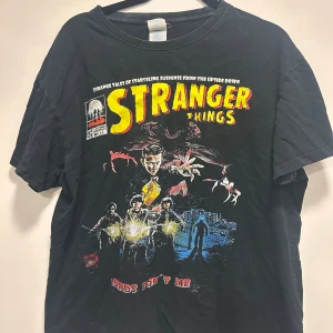 Svart Stranger Things t-shirt - Cool svart t-shirt med Stranger Things-tryck i rött och gult. Perfekt för fans av serien! Den har korta ärmar och är gjord i bomull, vilket gör den bekväm att bära. Passar bra till vardags eller för en avslappnad look.
