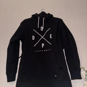 Svart hoodie från Dope - Säljer en svart snowboard jacka från Dope med vit text och grafiskt tryck på framsidan. Den har en klassisk känguruficka och justerbar huva med vita snören. 