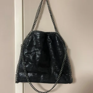 Svart axelväska med kedjedetaljer - Snygg svart axelväska i med coola kedjedetaljer runt kanterna och som axelrem, går att bära på två olika sett. Innerfick finns. Väskan har en glansig finish. Obetydligt använd 