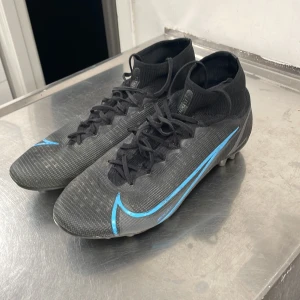 Svarta Nike mercurial fotbollsskor  - Säljer ett par svarta Nike fotbollsskor i bra skick. De har en snygg blå detalj på sidan och snörning för perfekt passform. Perfekta för fotbollsträning eller match. Skorna har en tight passform och är designade för att ge bra grepp på planen.