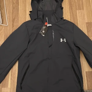 Svart jacka från Under Armour - Säljer en ny svart jacka från Under Armour i storlek M. Jackan har en justerbar huva och dragkedja framtill. Perfekt för kyliga dagar med sitt stilrena och funktionella design. Den är i syntetmaterial och har en sportig look.