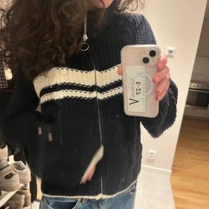 Marinblå stickad kofta med dragkedja - Säljer en mysig svart stickad kofta med dragkedja och vita ränder över bröstet. Den har en hög krage och är perfekt för kyliga dagar. Koftan är i bra skick är knappt använd 