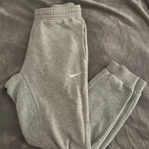 Grå mjukisbyxor från Nike - Säljer ett par grå mjukisbyxor från Nike i storlek S. De är superbekväma med elastisk midja och snörning. Perfekta för chill dagar eller träning. Byxorna har en liten Nike-logga på sidan och en bakficka.