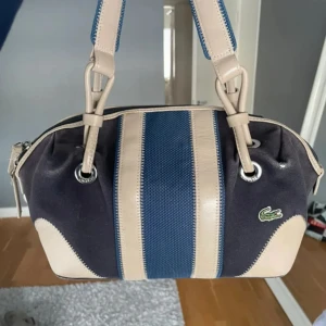 Lacoste Väska - Snygg axelväska från Lacoste i blått och beige med randigt mönster. Väskan har en dragkedja upptill och är perfekt för både vardag och fest. Insidan är randig i grönt och vitt, vilket ger en fräsch känsla. Den är i mycket bra skick och redo för nya äventyr!