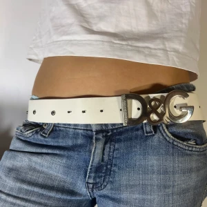 Vitt bälte från D&G - Snyggt vitt skinnbälte från Dolce & Gabbana med en stor silverfärgad D&G-spänne. Perfekt för att ge din outfit en lyxig touch. Bältet har små silverdetaljer längs hela längden. Passar perfekt till jeans för en stilren look. Använd det på fest eller till vardags för att sticka ut! 💞 125 cm