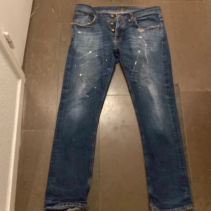 Blå jeans från Nudie Jeans Co - Säljer ett par blå jeans från Nudie Jeans i bra skick med en custom sliten design med färg fläckar på låren som är en del av designen på byxan