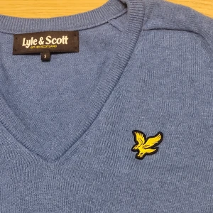 Blå tröja från Lyle & Scott - Säljer en snygg blå tröja från Lyle & Scott i storlek S. Tröjan har en v-ringning och ett gult broderat emblem på bröstet. Perfekt för både vardag och lite finare tillfällen. Den är i mycket bra skick och superbekväm att ha på sig.