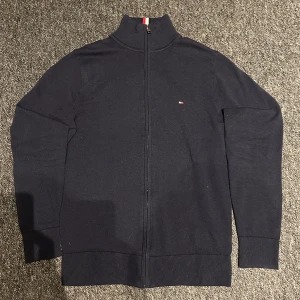 Stickad cardigan  - Säljer en stilren mörkblå stickad cardigan från Tommy Hilfiger i premium bomull. Den har en dragkedja framtill och ribbade muddar. Perfekt för höst och vinter, med en liten logga på bröstet och snygga detaljer i kragen. Passar både till vardags och lite finare tillfällen. Skicket är riktigt bra! Inga tecken på användning!! Stl Xs, nypris 1500 mitt pris 500kr
