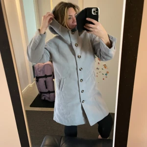 Ljusgrå kappa med knappar - Säljer en stilren ljusgrå kappa helt ny. Den har stora knappar framtill och en hög krage som skyddar mot kyla. Perfekt för höst och vår! Kappan har långa ärmar och en skön passform. Passar både till vardags och lite finare tillfällen.