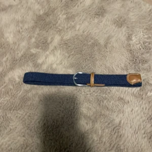 Blått flätat bälte med läderdetaljer - Säljer ett snyggt blått flätat bälte med silverfärgat spänne och bruna läderdetaljer. Perfekt för att ge en stilren touch till din outfit. Bältet är i mycket bra skick och passar både till vardags och fest. Justerbar längd för bästa passform.