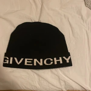 Svart mössa från Givenchy - Snygg svart mössa från Givenchy med vit logga. Perfekt för att hålla sig varm och stilren under kyliga dagar. Passar till alla outfits och ger en lyxig touch. Ett måste för modeälskare!