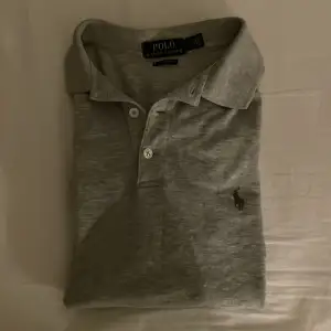 Säljer en stilren grå pikétröja från Ralph Lauren i slim fit. Tröjan har korta ärmar och klassisk krage med två knappar. Perfekt för både vardag och festliga tillfällen. Den är använd ett fåtal gånger, är i väldigt bra skick och redo för en ny ägare!