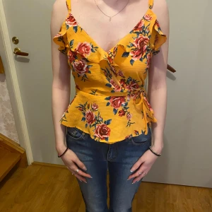 Gul blommig omlottopp - Säljer en superfin gul omlottopp med blommigt mönster. Toppen har volangdetaljer och smala axelband, perfekt för sommardagar. Den är figursydd och ger en snygg siluett. Passar både till jeans och kjol för en fräsch look💛