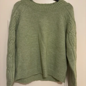 Grön stickad tröja från Vero Moda - Mysig grön stickad tröja från Vero Moda med kabelstickade detaljer på ärmarna. Tröjan har en rund halsringning och ribbade muddar vid ärmslut och nederkant. Perfekt för kyliga dagar!🙌🏼🤍