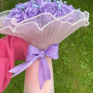 Lila ros-bukett i satin - En vacker bukett med lila rosor och små blå blommor, insvept i ett vitt spetsliknande papper med pärldetaljer. Perfekt som en romantisk gåva eller dekoration. OBS: beställs minst 10 dagar innan leverans 🚚