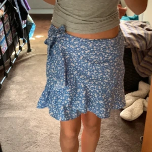 Blå blommig omlottkjol - Söt blå omlottkjol med vita blommor och volangdetaljer. Kjolen har en knytning i midjan och är perfekt för varma dagar.