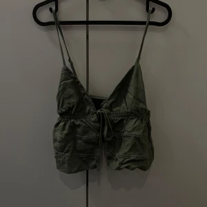 Olivgrön linnetopp med spetsdetaljer - Snygg olivgrön linnetopp med smala axelband och spetsdetaljer. Toppen har en knytning framtill och en luftig passform, perfekt för varma dagar.