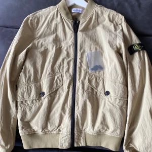 Beige bomberjacka från Stone Island - Snygg beige bomberjacka från Stone Island med klassisk logga på ärmen. Jackan har dragkedja framtill och två fickor med knappar. Perfekt för en stilren look. Den är aldrig använd och är i storleken 170 motsvarar S/small. Fick den i julklapp men den var tyvärr för liten, original priset på jackan ligger runt 3999 kr.