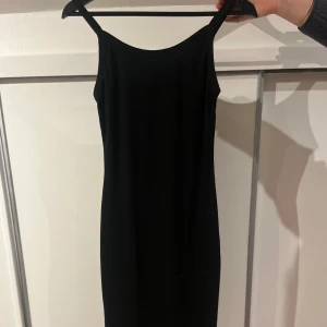 Svart ärmlös klänning - Elegant svart klänning med en tight passform. Den är ärmlös och har en klassisk design som passar perfekt för festliga tillfällen. Klänningen är gjord av ett mjukt och bekvämt material. Ca knälång. 