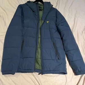Blå pufferjacka med huva - Snygg blå pufferjacka från Lyle & Scott med huva och dragkedja. Jackan har ett litet gult broderat emblem på bröstet och praktiska fickor med dragkedja. Perfekt för kyliga dagar.