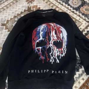 Svart tröja med dödskalle från Philipp Plein - Snygg svart tröja från Philipp Plein med ett stort dödskallemotiv i rött, vitt och blått på framsidan. Tröjan har långa ärmar och en rund halsringning. Perfekt för en cool och avslappnad stil.