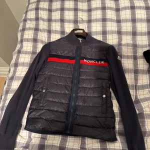 Mörkblå jacka från Moncler - Snygg mörkblå cardigan från Moncler med röd och vit logga på bröstet. Det är 2020 års modellen, Cardiganen har en quiltad design framtill och dragkedja. Den är långärmad med ribbade muddar och krage. Den är riktigt skön och används oftast som kofta eller tröja under jacka. Pris kan diskuteras, bara att höra av dig vid några funderingar!🤩