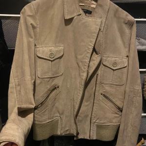 Beige mockajacka från KappAhl - Snygg beige mockajacka från KappAhl i storlek S. Jackan har långa ärmar, dragkedja och knappar framtill samt praktiska fickor med lock. Perfekt för en stilren look. Has a small rip on collar.