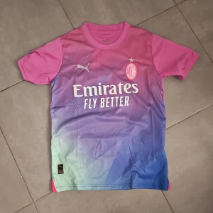 AC Milan fotbollströja från Puma - Snygg AC Milan fotbollströja från Puma i en cool färgövergång från rosa till blå och grön. Tröjan har korta ärmar och tryck med 'Emirates Fly Better' och 'wefox'. Perfekt för fans som vill sticka ut! Hör av er vid intresse!:)