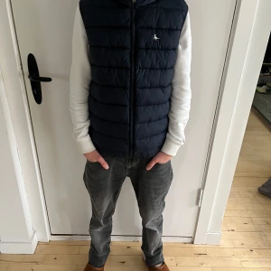 Mörkblå barn dunväst med dragkedja - Säljer en snygg mörkblå barn dunväst med dragkedja framtill. Västen har en liten logga på bröstet och är perfekt för kyliga dagar. Den är både stilren och praktisk, idealisk för höst och vår.