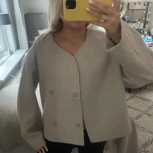 Beige kappa med knappar - Säljer en stilren beige kappa med dubbelknäppning och rund hals. Jackan har en lös passform och långa ärmar, perfekt för höst och vår. En tidlös design som passar till många olika outfits.