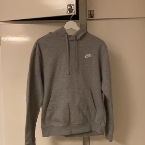 Grå hoodie från Nike - Säljer en klassisk grå hoodie från Nike med justerbar huva och känguruficka. Perfekt för en avslappnad stil. Nike-loggan är tryckt i vitt på bröstet.