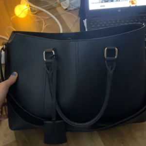 Svart axelväska med gulddetaljer - Snygg svart axelväska i skinn med eleganta gulddetaljer. Väskan har en rymlig insida och praktiska handtag för enkel bärning. Perfekt för att bära med sig det nödvändigaste med stil.