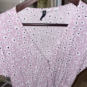 Ljuslila blommig klänning - Söt ljuslila klänning med blommigt mönster i vitt och svart. Klänningen har en v-ringad design och knytband i midjan för en smickrande passform. Perfekt för vår och sommar!