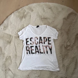 Vit t-shirt med tryck från Gina Tricot - Säljer en vit t-shirt från Gina Tricot med texten 'ESCAPE REALITY' i svart och rosa på framsidan. T-shirten har en v-ringad hals och korta ärmar. Perfekt för en avslappnad stil.