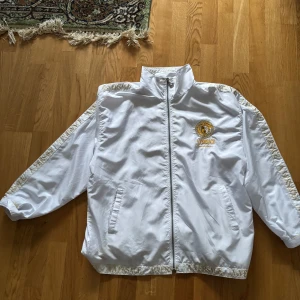 Vit jacka med gulddetaljer - Snygg vit jacka med dragkedja och gulddetaljer från Aversace. Jackan har ett broderat emblem på bröstet och ryggen, samt text längs kragen. Perfekt för en stilren look.