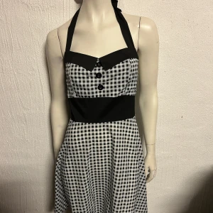 Svartvit rutig klänning - Charmig svartvit rutig klänning med halterneck och knappar framtill. Den har en markerad midja och en klassisk 50-talsinspirerad stil. Perfekt för en retro-look!