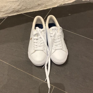 Vita sneakers från Polo - Snygga vita sneakers från Polo med klassisk design och snörning. Skorna har en broderad logotyp på sidan och texten 'POLO' på hälen. Perfekta för en stilren look.