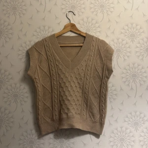 Beige stickad väst - Snygg beige stickad väst med v-ringning och kabelstickat mönster. Perfekt för lager-på-lager under höst och vinter.