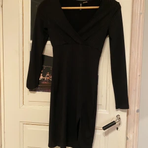 Svart ribbad klänning - Snygg svart ribbad klänning med v-ringad hals och långa ärmar. Klänningen har en elegant slits framtill och omlott-design som ger en stilren look. Perfekt för både vardag och festliga tillfällen.