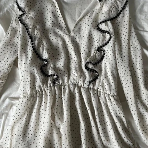 Vit prickig klänning från Zara -  vit klänning från Zara med svarta prickar och volangdetaljer. Klänningen har en v-ringad hals och långa ärmar. Perfekt vid finare tillfällen ❣️💓
