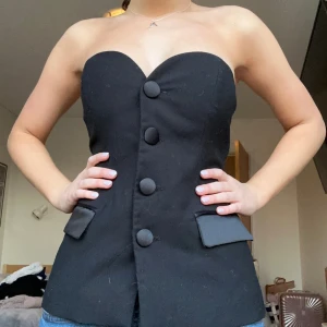 Svart axelbandslös korsettopp - Snygg svart axelbandslös korsettopp med stora knappar framtill. Toppen har en figurnära passform och dekorativa fickdetaljer. Perfekt för en stilren look.💓använt max 3 gånger!!