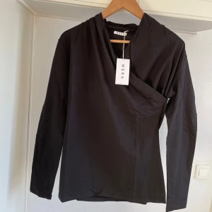 Svart långärmad topp från Wera M - Snygg svart långärmad topp från Wera med en unik omlott-design vid halsen. Perfekt för en stilren look. Toppen är i ett mjukt och bekvämt material.