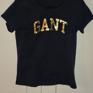Mörkblå t-shirt från GANT - Snygg svart t-shirt från GANT med korta ärmar och ett guldtryck av märkesnamnet. M i storlek men passar även S